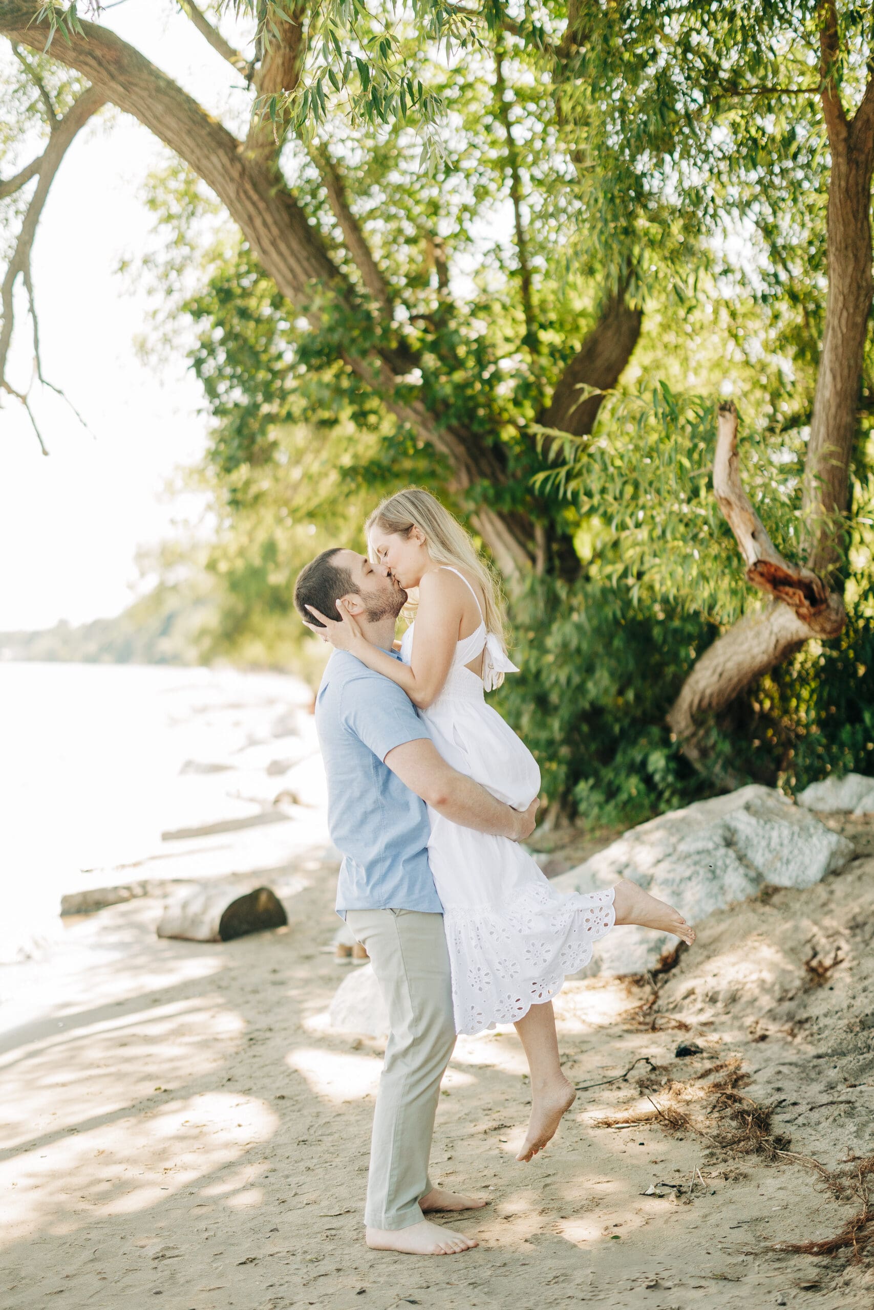 Couple laughing as the man lifts the woman under a canopy of trees by the lake.