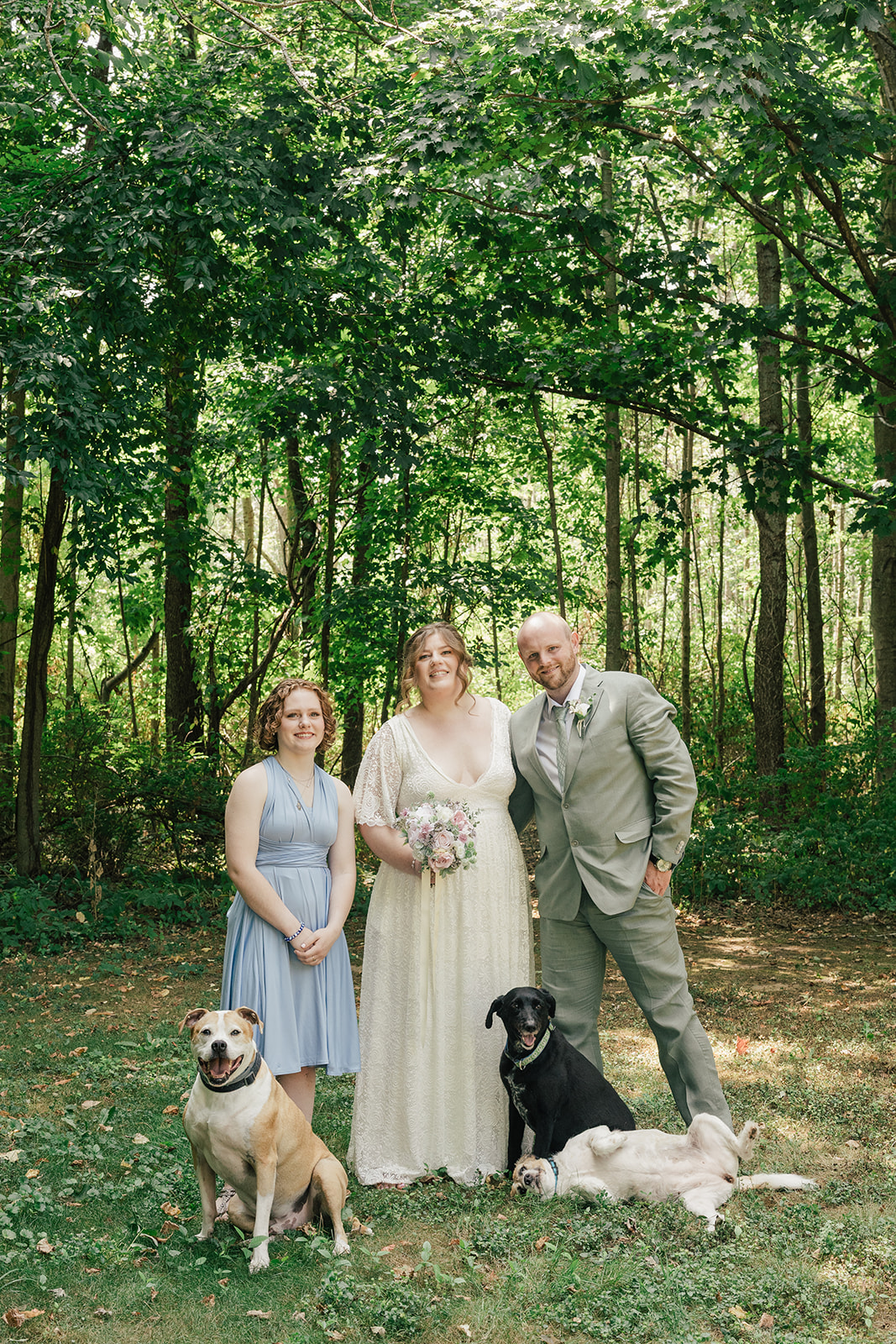 A bride and groom standing with their daughter and three dogs in a wooded outdoor setting. The bride holds a bouquet and the groom leans in close, while the dogs sit and play in the grass in front of them.