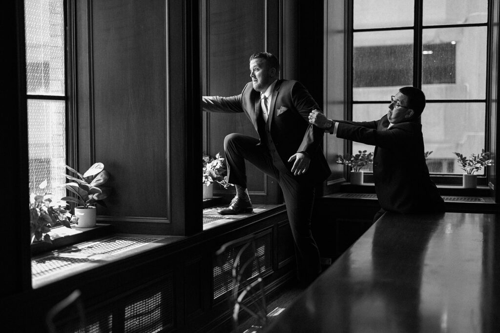 Rochester wedding photographer capturing a funny documentary moment of a groom pretending to jump from a window while the father of the bride holds him back.